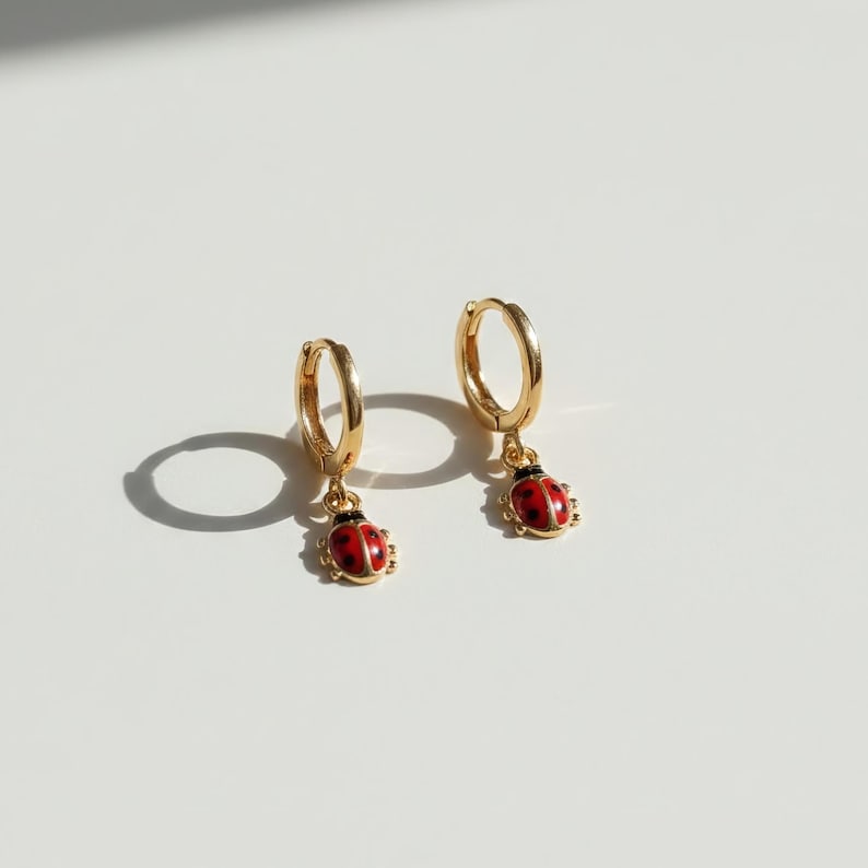 May include: Two gold-coloured hoop earrings, each with a small, red ladybird charm. The ladybirds have black spots and hang from the hoops. The earrings are shown on a white surface.