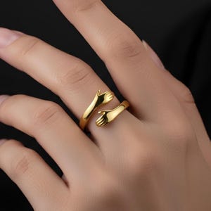 May include: A gold-colored ring featuring a hugging hands design. The ring wraps around the finger with two hands meeting. The ring is made of a shiny, reflective metal.