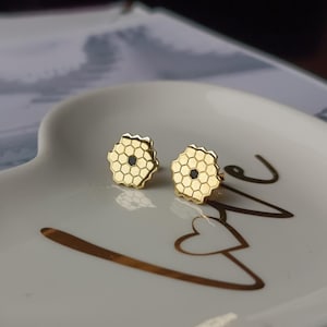 May include: A pair of gold-colored stud earrings. Each earring features a honeycomb design with a dark center. The earrings are displayed on a white heart-shaped dish with gold lettering.