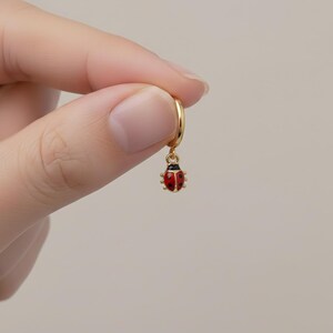 May include: A gold hoop earring featuring a ladybug charm. The ladybug is red with black spots and a black head. The earring is suspended from the hoop. The earring is held by a person's fingers against a plain background.