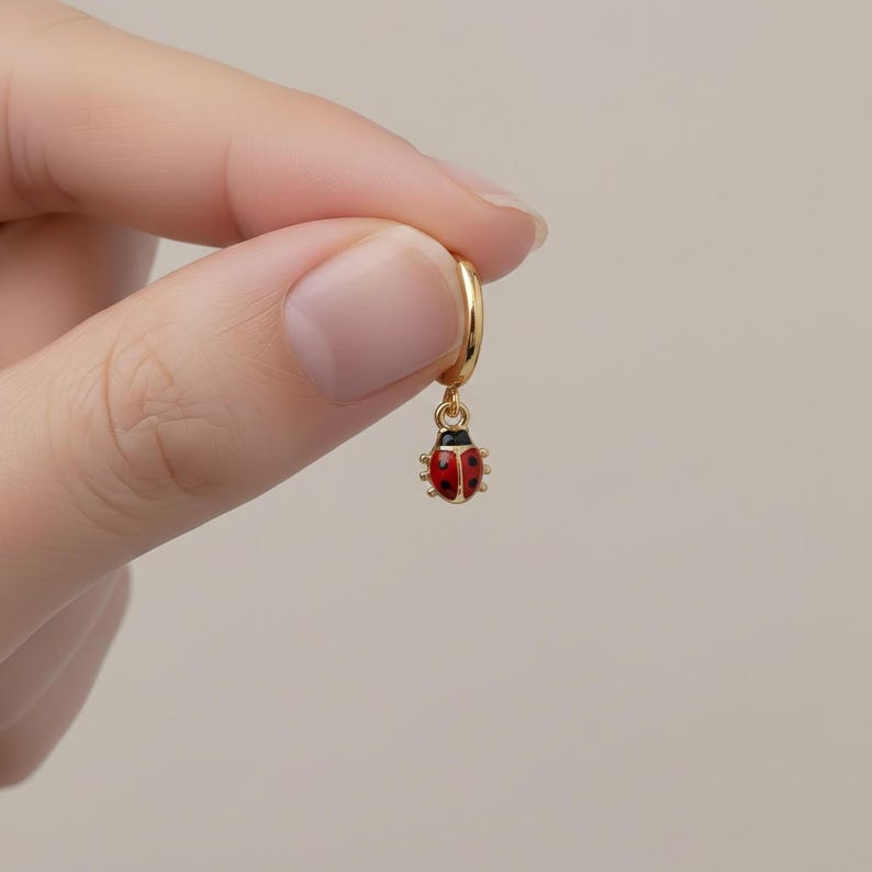 May include: A gold hoop earring featuring a ladybug charm. The ladybug is red with black spots and a black head. The earring is suspended from the hoop. The earring is held by a person's fingers against a plain background.