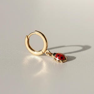 May include: A gold-coloured hoop earring featuring a ladybug charm. The ladybug charm is red with black spots and a black head. The earring is made of a shiny, metallic material.
