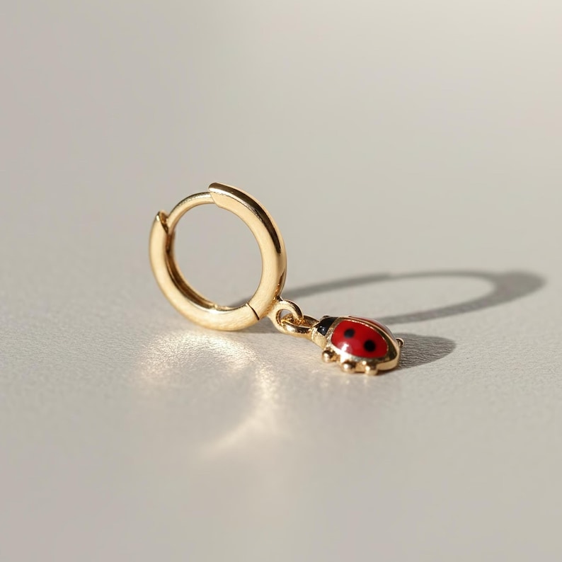 May include: A gold-coloured hoop earring featuring a ladybug charm. The ladybug charm is red with black spots and a black head. The earring is made of a shiny, metallic material.