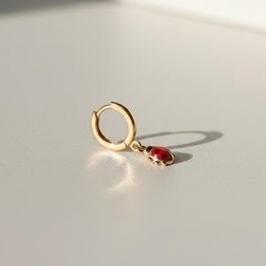 May include: A gold-coloured hoop earring featuring a ladybird charm. The ladybird is red with black spots and dangles from a small chain. The earring is on a white surface with a shadow.