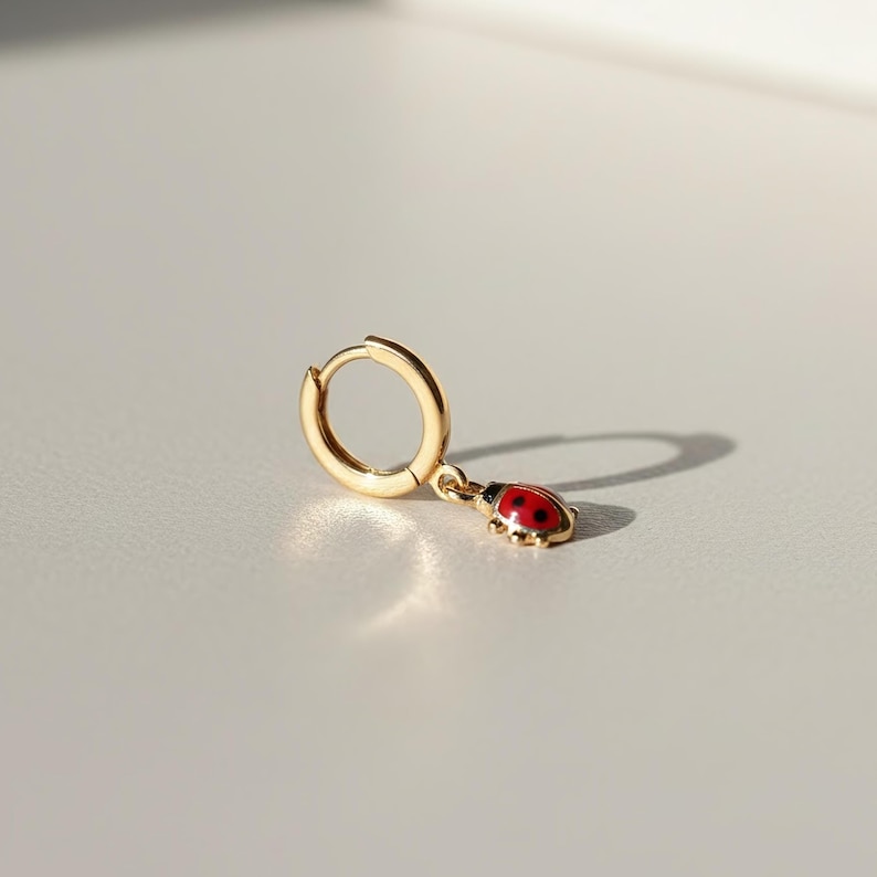 May include: A gold-coloured hoop earring featuring a ladybird charm. The ladybird is red with black spots and dangles from a small chain. The earring is on a white surface with a shadow.