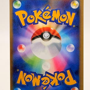 以下が含まれることがあります： 金色の縁取りと青い背景のポケモンカード。 「ポケモン」の文字は黄色とオレンジ色で、中央に赤と白のモンスターボールがあります。 さまざまな色のオーブと稲妻も見えます。