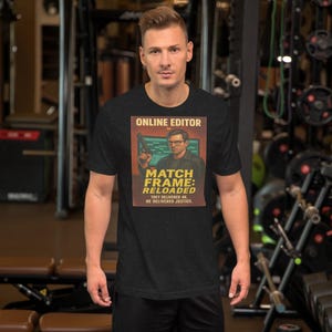 Puede incluir: Una camiseta gris oscuro con un gráfico que dice "ONLINE EDITOR" y "MATCH FRAME: RELOADED". El gráfico muestra a un hombre sosteniendo un arma. La camiseta la lleva una persona en un gimnasio.