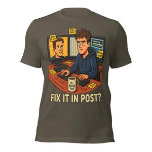Op de afbeelding: Olijfgroen t-shirt met een cartoonillustratie van een persoon achter een computer met de tekst "FIX IT IN POST?". De illustratie bevat een koffiemok met de tekst "It's Already Post". Het ontwerp bevat ook twee computermonitoren en plakbriefjes.