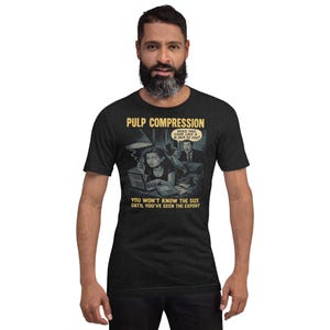May include: Dark gray t-shirt with the text "PULP COMPRESSION" and a comic-style illustration. The illustration features a woman at a laptop and a man in a suit. The text "YOU WON'T KNOW THE SIZE UNTIL YOU'VE SEEN THE EXPORT" is also visible.