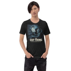 May include: Dark heather gray t-shirt featuring a graphic of a person looking at a document on a computer screen, with the text "THE CUT THING" and "The ultimate impossible notes terror".