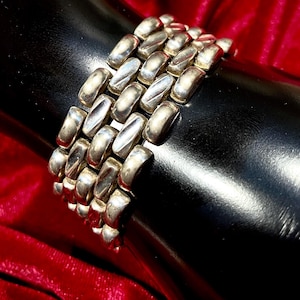 May include: Silver-toned bracelet with a linked design. The bracelet is composed of multiple rows of elongated, rectangular links. The links have a slightly curved shape and a polished finish. The bracelet is displayed against a black surface and a red velvet background.