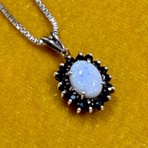 May include: A silver pendant necklace featuring a central oval-shaped iridescent white opal stone surrounded by a halo of small, dark blue faceted gemstones. The pendant hangs from a delicate silver box chain with a polished metallic finish.