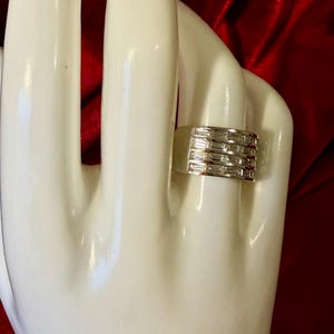 May include: A silver ring with a rectangular design. The ring is wide and features multiple rows of rectangular-cut stones. The ring is displayed on a white hand against a red background.