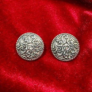 May include: Two silver-toned, round earrings with intricate, swirling floral patterns. Each earring has a rope-like border. The earrings are displayed on a red velvet surface, creating a striking contrast.