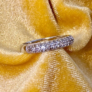 May include: A silver ring with a band encrusted with small, sparkling stones. The ring is set against a backdrop of a golden-yellow, textured fabric, creating a contrast of cool metal and warm color. The ring appears to be a piece of jewelry.