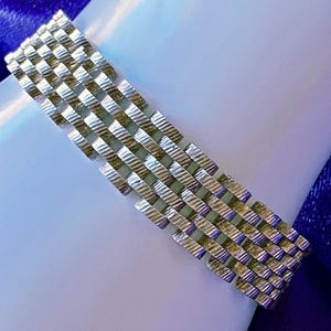 May include: A silver-toned bracelet with a woven design. The bracelet is rectangular and features a series of interconnected, textured metal bars. The bracelet is displayed on a white surface with a blue background.