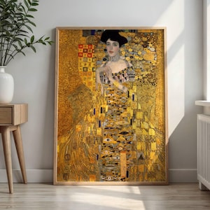 May include: A framed print of a Gustav Klimt painting. The artwork features a woman in a golden dress and hat, with intricate patterns and details. The color palette is dominated by gold, yellow, and black.