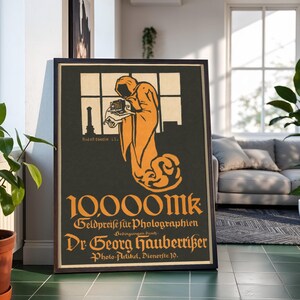 May include: A framed vintage poster featuring a hooded figure in orange holding a camera against a black background. The text reads 10,000 Mk, Geldpreise für Photographien, Bedingungen durch Dr. Georg Hauberrisser, and Photo-Artikel, Dienerstr. 10.