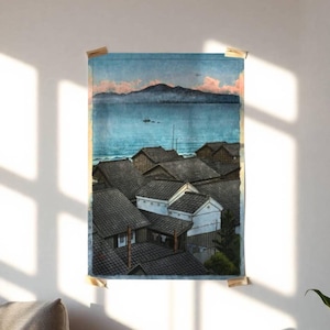 May include: A framed print depicting a coastal village scene. The artwork features a view of rooftops, a body of water, and mountains in the distance. The color palette includes shades of blue, gray, and brown, with hints of pink in the sky. The print is attached to the wall with wooden clips.