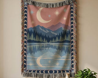 May include: A woven throw blanket featuring a landscape scene with a crescent moon, stars, mountains, and a forest reflected in a lake. The border displays a repeating moon phase pattern in dark blue and cream, finished with a fringed edge.