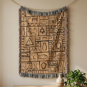 May include: A rectangular woven throw blanket featuring a tan background with a grid of black hand-drawn style symbols, including a fish, a star, a bottle, and the letters BNI. The blanket has a fringed edge with multicolored threads.