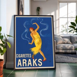 May include: A framed vintage-style poster featuring a person in a yellow dress dancing against a deep blue background, with white smoke swirls and the text CIGARETTES ARAKS printed in large white letters at the bottom of the rectangular print.