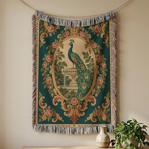 May include: A woven throw blanket featuring a central oval medallion with a peacock perched on a stone fountain. The design is surrounded by a border of pink roses and scrolling foliage on a deep teal background, finished with decorative fringe.