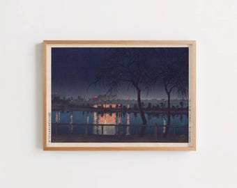 May include: A framed woodblock print depicting a night scene with dark blue skies, silhouettes of bare trees, and a calm body of water reflecting orange city lights. A dark fence runs across the foreground, and Japanese text is visible on the sides.