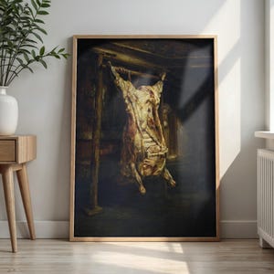 May include: A framed wall art print featuring a dark, moody oil painting of a butchered animal carcass hanging from a wooden frame structure. The artwork displays deep shadows, warm golden highlights on the meat, and a somber, textured aesthetic.