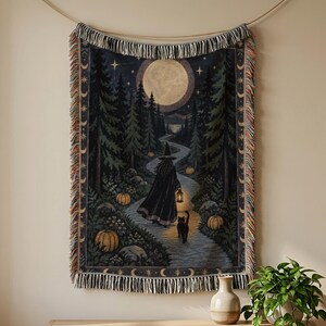 May include: A woven throw blanket featuring a dark forest scene with a full moon, a figure in a black cloak and pointed hat holding a lantern, and a black cat walking on a path lined with orange pumpkins, finished with decorative fringe edges.