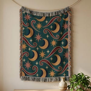 May include: A woven throw blanket featuring a dark teal background with a repeating pattern of gold crescent moons, multi-pointed stars in shades of pink and yellow, and swirling maroon lines. The blanket has a decorative fringe on all sides.