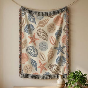 May include: A woven throw blanket featuring a repeating pattern of seashells, starfish, and sand dollars in shades of blue, tan, and coral on a cream background. The blanket has decorative fringe along the top and bottom edges with multicoloured threads.