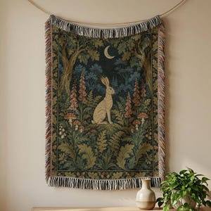 May include: A woven tapestry throw blanket featuring a central light brown hare sitting in a dark forest setting with tall pink foxglove flowers, red and white mushrooms, ferns, and a crescent moon in the night sky, finished with fringed edges.