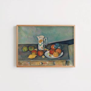 May include: A framed still life painting depicting a white ceramic pitcher with a floral pattern placed on a wooden table. A white plate holds several round fruits including oranges and apples, while other loose fruits are scattered on the surface.