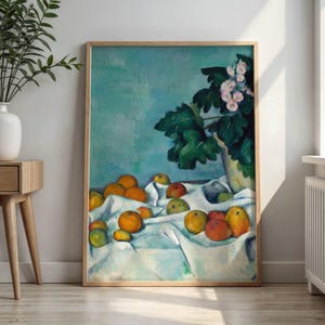 May include: A framed art print featuring a still life painting of various round fruits in shades of orange, yellow, and green resting on a white draped cloth. A vase with dark green leaves and light pink flowers sits to the right against a teal background.