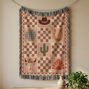 May include: A woven throw blanket featuring a grid pattern of pink and cream squares alternating with icons including a brown cowboy hat, a disco ball, a horseshoe, a green cactus, a floral cow skull, and a sunburst, finished with fringed edges.