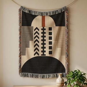 May include: A woven throw blanket featuring a geometric pattern in black, beige, and rust orange. The design includes a central arch, vertical columns of chevrons, squares, and rectangles, with multi-colored fringe along the vertical edges.