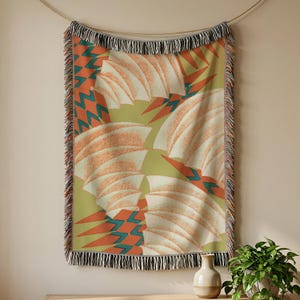 May include: A woven throw blanket featuring a repeating pattern of cream-colored fan shapes on a muted olive green background, accented by orange and teal geometric zig-zag motifs. The blanket has a decorative fringe along the top and bottom edges.