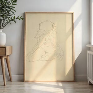 May include: A framed pencil sketch depicting a seated figure, drawn with fine lines. The artwork, set against a cream background, is enclosed in a light-toned wooden frame. The sketch captures the form of a person in a seated position.