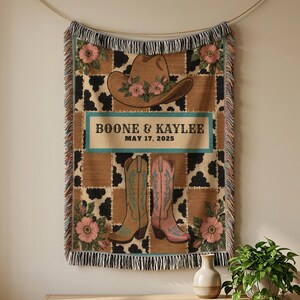 May include: A woven throw blanket featuring a brown cowboy hat with pink flowers, a pair of cowboy boots, and a black and tan cow print pattern. The text BOONE & KAYLEE and MAY 17, 2025 is centered on a teal rectangular background with fringe edges.