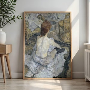 May include: A framed print of a painting depicting a woman seated, viewed from the back. She has red hair pulled back, and is draped in white fabric. The artwork has a muted color palette with shades of beige, gray, and brown. The frame is a light wood color.