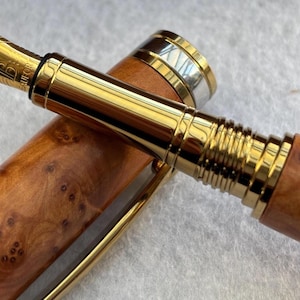 Puede incluir: Una pluma estilogr&aacute;fica de lujo con plum&iacute;n y detalles dorados. El cuerpo de la pluma es de madera marr&oacute;n claro con un patr&oacute;n de vetas &uacute;nico. El capuch&oacute;n es parcialmente visible, con bandas doradas y plateadas.