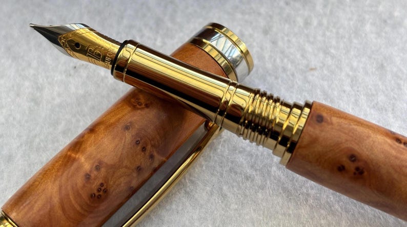 Puede incluir: Una pluma estilogr&aacute;fica de lujo con plum&iacute;n y detalles dorados. El cuerpo de la pluma es de madera marr&oacute;n claro con un patr&oacute;n de vetas &uacute;nico. El capuch&oacute;n es parcialmente visible, con bandas doradas y plateadas.