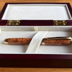 Puede incluir: Un bol&iacute;grafo de madera con un patr&oacute;n de madera de ra&iacute;z y detalles dorados descansa en una caja de presentaci&oacute;n de madera burdeos con un interior color crema. La caja est&aacute; abierta, revelando el bol&iacute;grafo y su elegante almacenamiento.