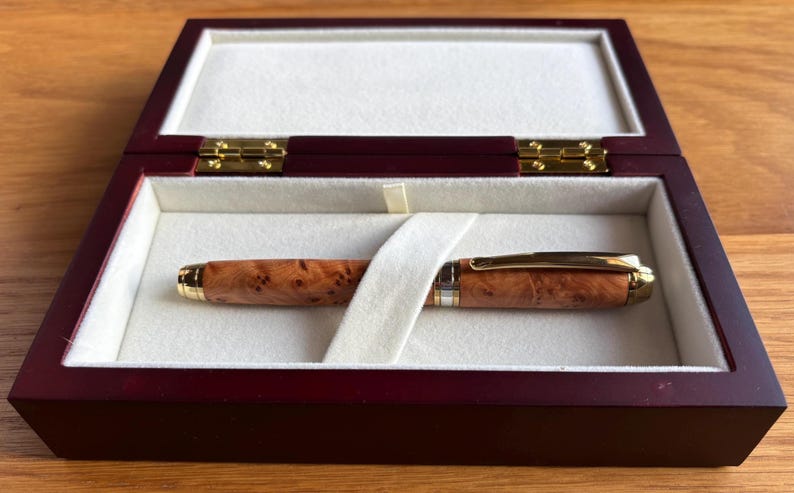 Puede incluir: Un bol&iacute;grafo de madera con un patr&oacute;n de madera de ra&iacute;z y detalles dorados descansa en una caja de presentaci&oacute;n de madera burdeos con un interior color crema. La caja est&aacute; abierta, revelando el bol&iacute;grafo y su elegante almacenamiento.