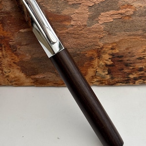 May include: A dark brown wooden pen with a silver clip and accents. The pen has a sleek, modern design and appears to be made of high-quality materials. The pen is approximately 14 cm long.
