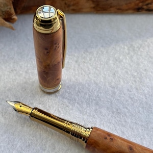 Puede incluir: Una pluma estilogr&aacute;fica artesanal con cuerpo de madera y detalles dorados. La pluma tiene un plum&iacute;n dorado y un capuch&oacute;n con clip dorado. El patr&oacute;n de la veta de la madera es visible en el cuerpo.