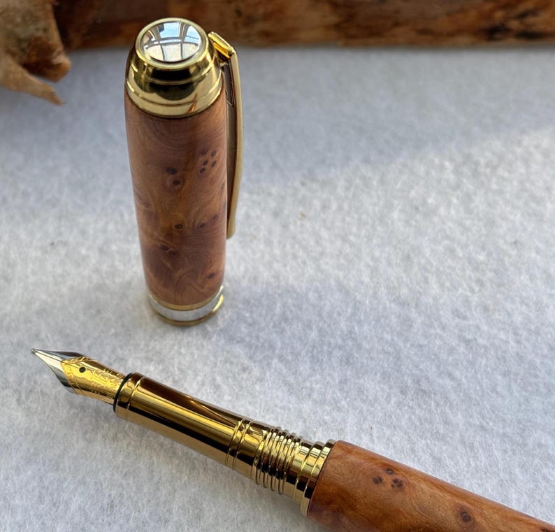 Puede incluir: Una pluma estilogr&aacute;fica artesanal con cuerpo de madera y detalles dorados. La pluma tiene un plum&iacute;n dorado y un capuch&oacute;n con clip dorado. El patr&oacute;n de la veta de la madera es visible en el cuerpo.