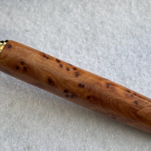 Puede incluir: Primer plano de un bol&iacute;grafo de madera con detalles dorados. El cuerpo del bol&iacute;grafo es de color marr&oacute;n claro con manchas marrones oscuras. La punta y el capuch&oacute;n del bol&iacute;grafo son dorados.