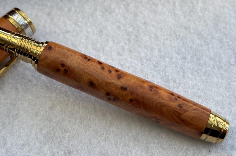 Puede incluir: Primer plano de un bol&iacute;grafo de madera con detalles dorados. El cuerpo del bol&iacute;grafo es de color marr&oacute;n claro con manchas marrones oscuras. La punta y el capuch&oacute;n del bol&iacute;grafo son dorados.